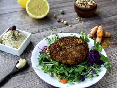 Hamburguesa vegetal dorada sobre brotes verdes, con limón y legumbres al fondo