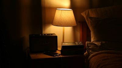 Mesa de noche con una lámpara de luz cálida junto a la cama en un cuarto oscuro, un ambiente que ayuda a entender para qué sirve la melatonina