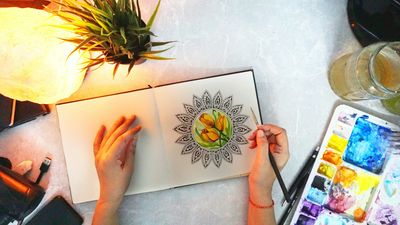 Manos dibujando un mandala en un cuaderno junto a acuarelas, uno de los símbolos espirituales más conocidos